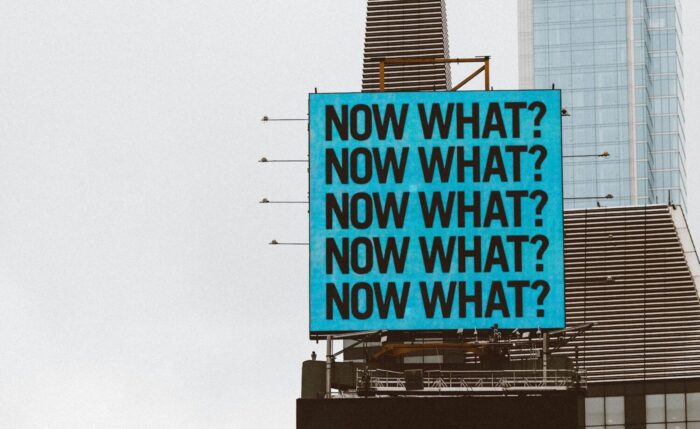 a large blue sign on top of a tall building "Now What?"